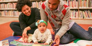 A man and woman smile at a toddler reading a book on the floor
