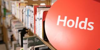 Photograph of a library shelf with a red circular sign with white text that reads: Holds