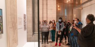 A group of people in face masks stand to the right of a display case while a woman in the foreground gestures towards the cases and the items on display.