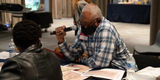 A man in a plaid shirt and a black face mask sits at a table while holding a microphone and looking down at a historical map. 