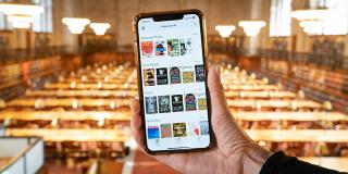 Photo of a hand holding an iPhone in front of a room full of tables and chandeliers; the iPhone has SimplyE on the screen.