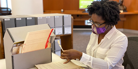 A woman wearing a purple, black, and white face mask is sitting at a table and reading papers from the Schomburg Center’s collections. 